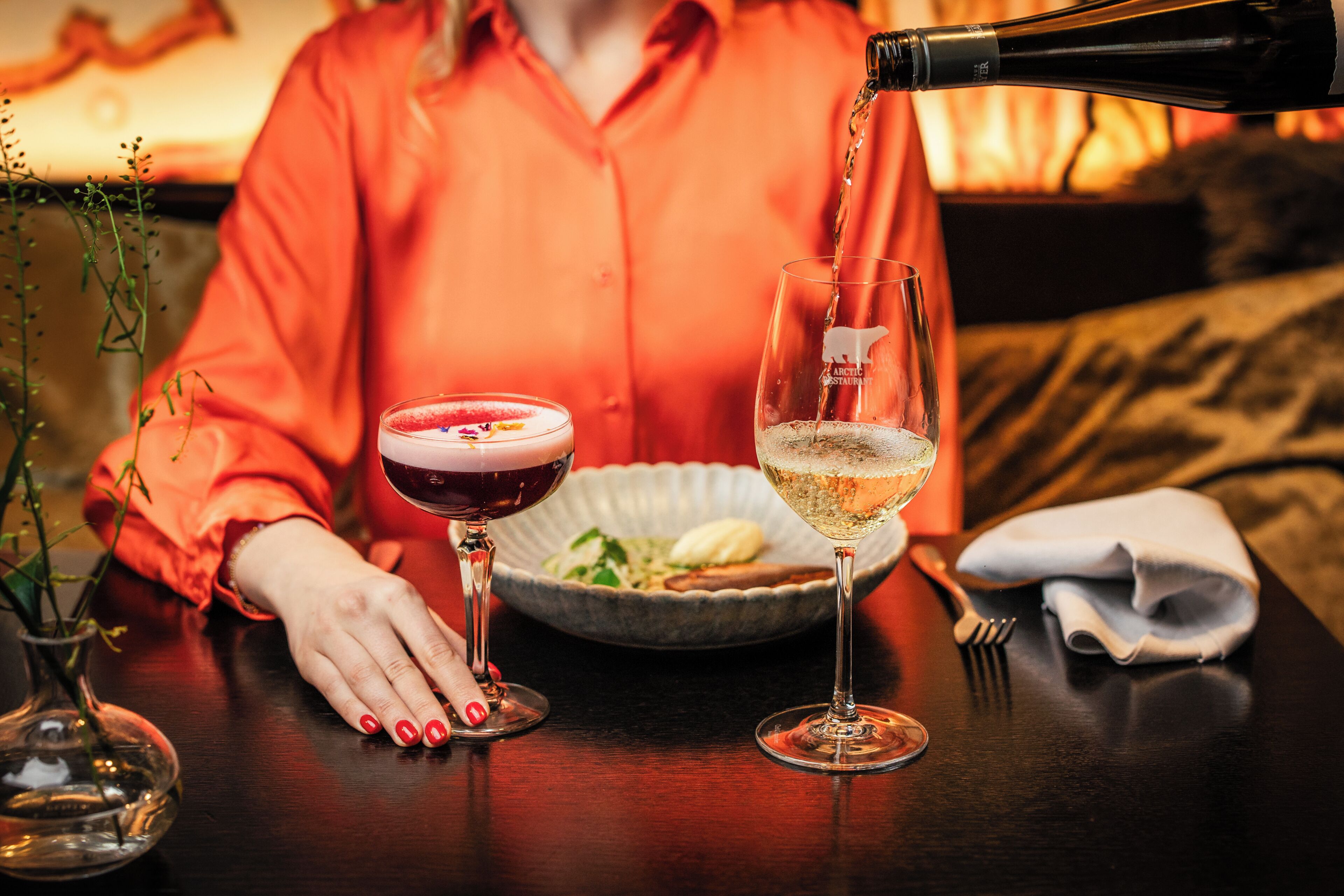 Une table avec deux verres, l’un rempli d’une boisson rouge et l’autre d’un liquide transparent, une bouteille versant son contenu dans le verre transparent, et un bol de nourriture.