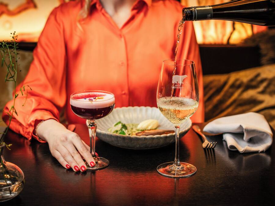 Une table avec deux verres, l’un rempli d’une boisson rouge et l’autre d’un liquide transparent, une bouteille versant son contenu dans le verre transparent, et un bol de nourriture.