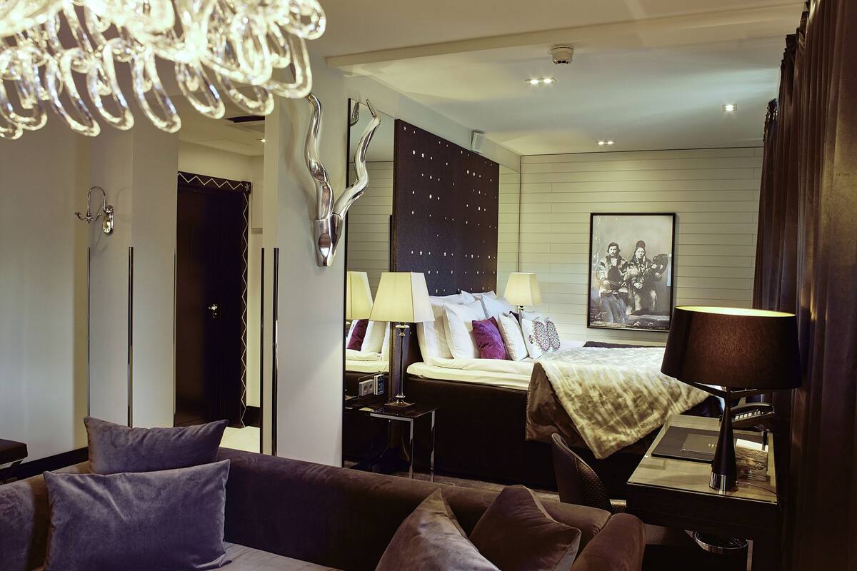 A modern hotel room with a large bed, a sitting area with a sofa, a desk with a lamp, a large mirror, and a chandelier.