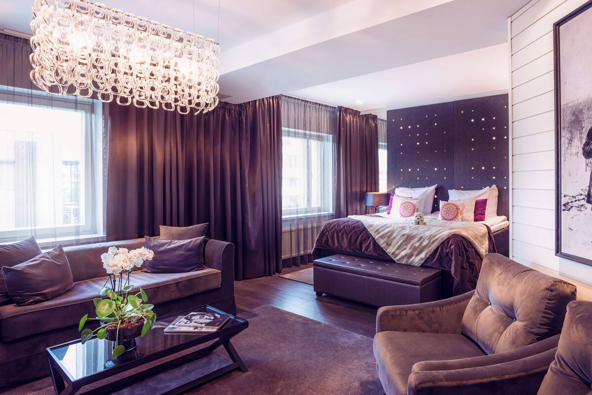 A modern hotel room with a large bed, a sofa, a coffee table, and a chandelier.