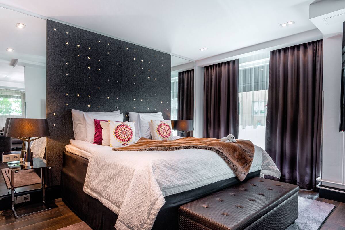 A modern bedroom with a large bed, a bedside table with a lamp, a flat-screen TV, and a large window with curtains.