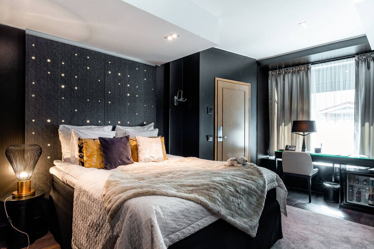 A modern bedroom with a star-patterned wall, a bed with multiple pillows, a bedside table with a lamp, and a desk with a chair.