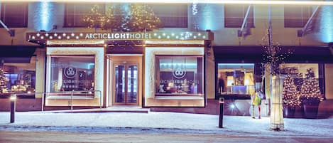 The exterior of the Arctic Light Hotel with a snow-covered ground, illuminated entrance, and a Christmas tree decoration.