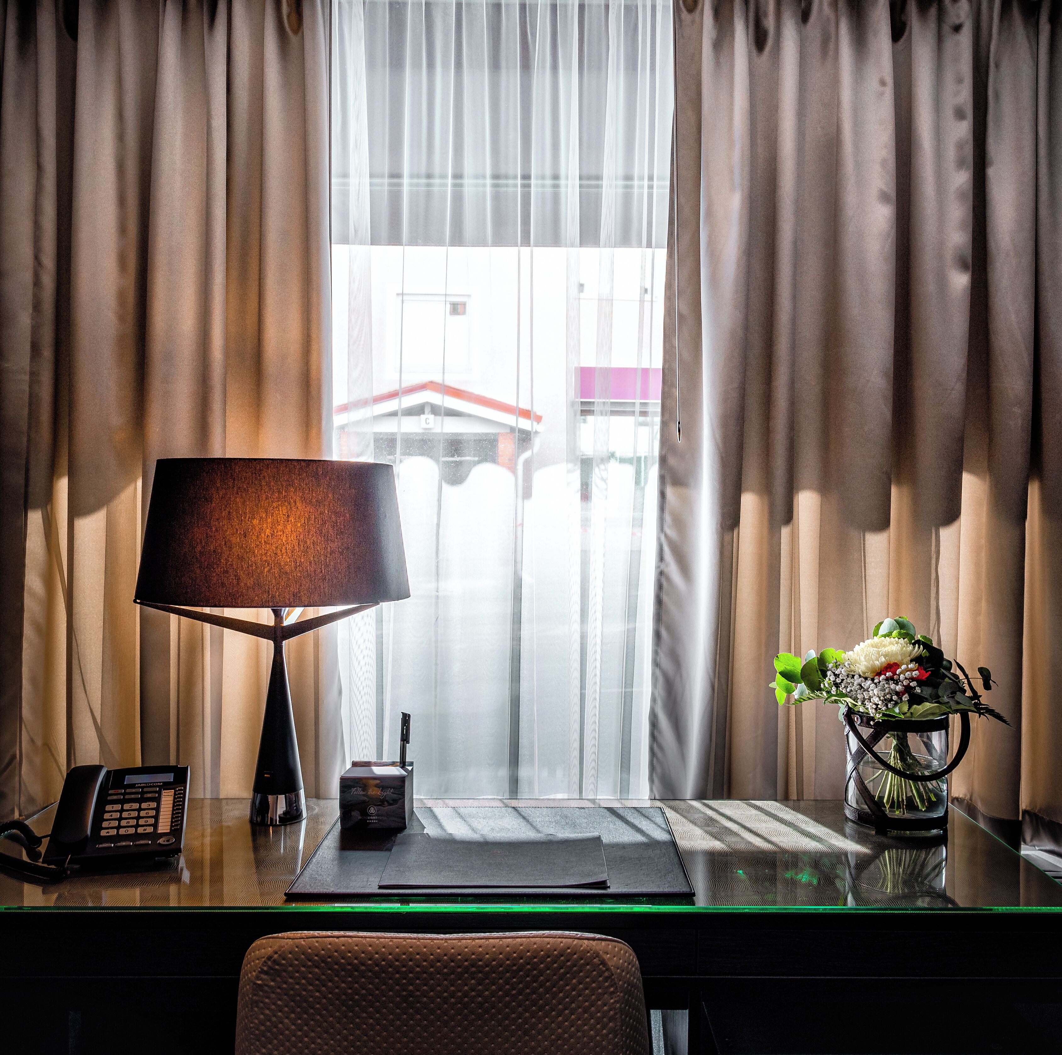 Une chambre d’hôtel équipée d’un bureau, d’une lampe, d’un téléphone, d’un vase avec des fleurs et offrant une vue sur un immeuble à travers des rideaux transparents.