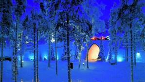 Escena nevada de un bosque con tiendas tipi iluminadas y una estructura brillante y cálida al fondo.