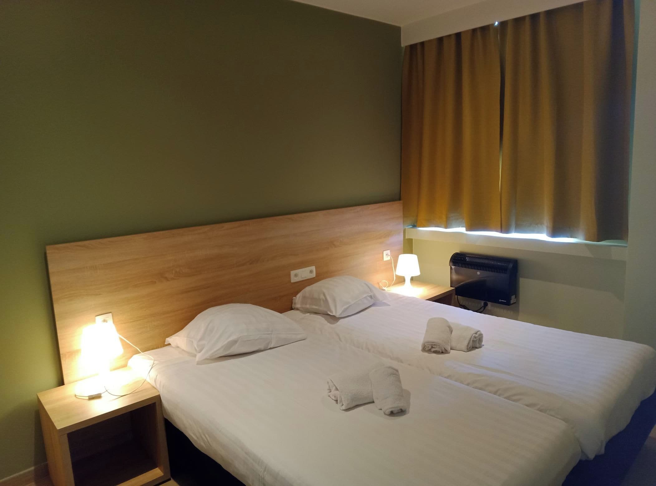 A hotel room with a wooden headboard, two bedside tables with lamps, a bed with white linens, and a window with curtains.