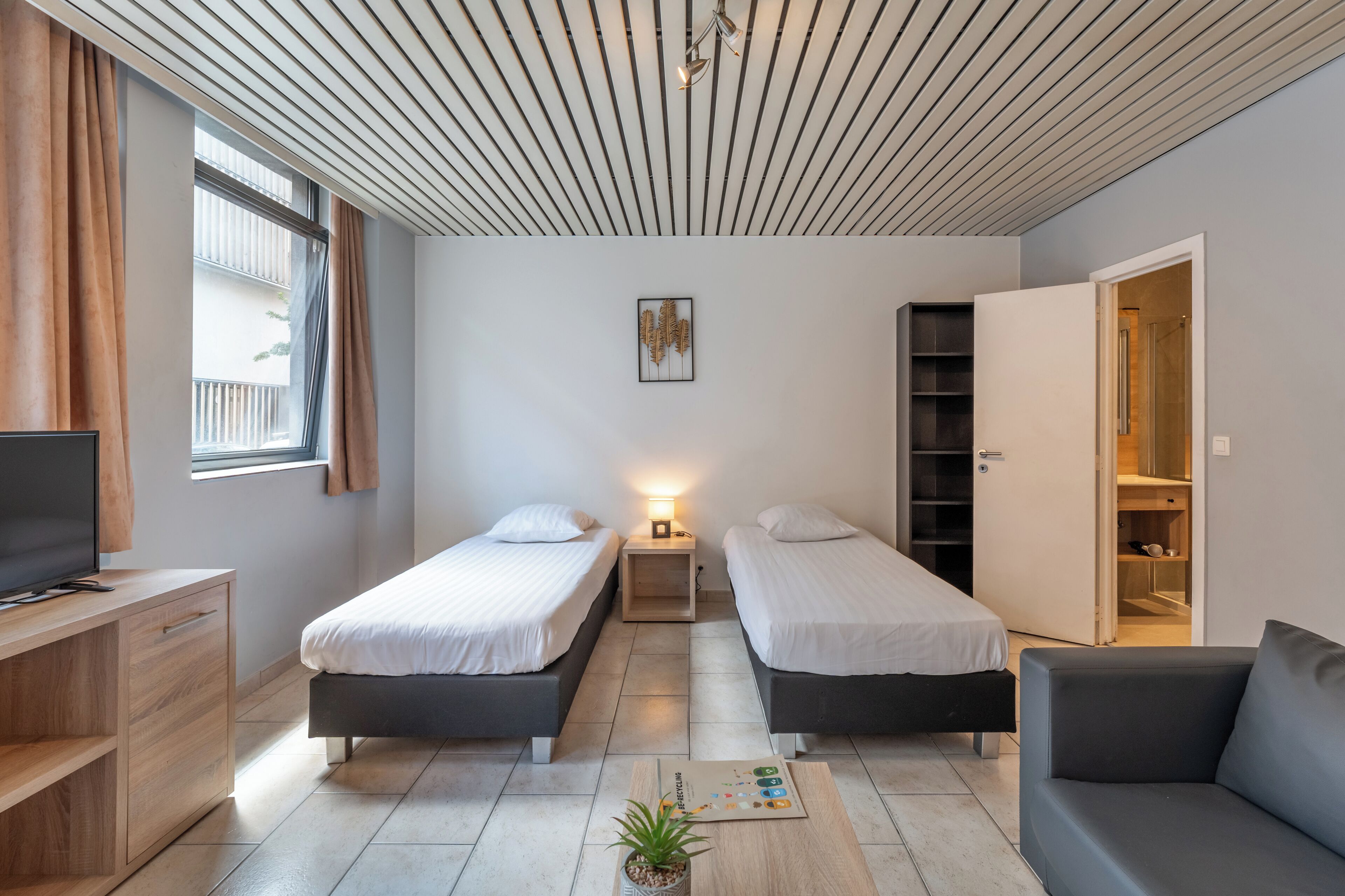 A modern bedroom with two single beds, a desk, a TV, a bookshelf, and a wardrobe.