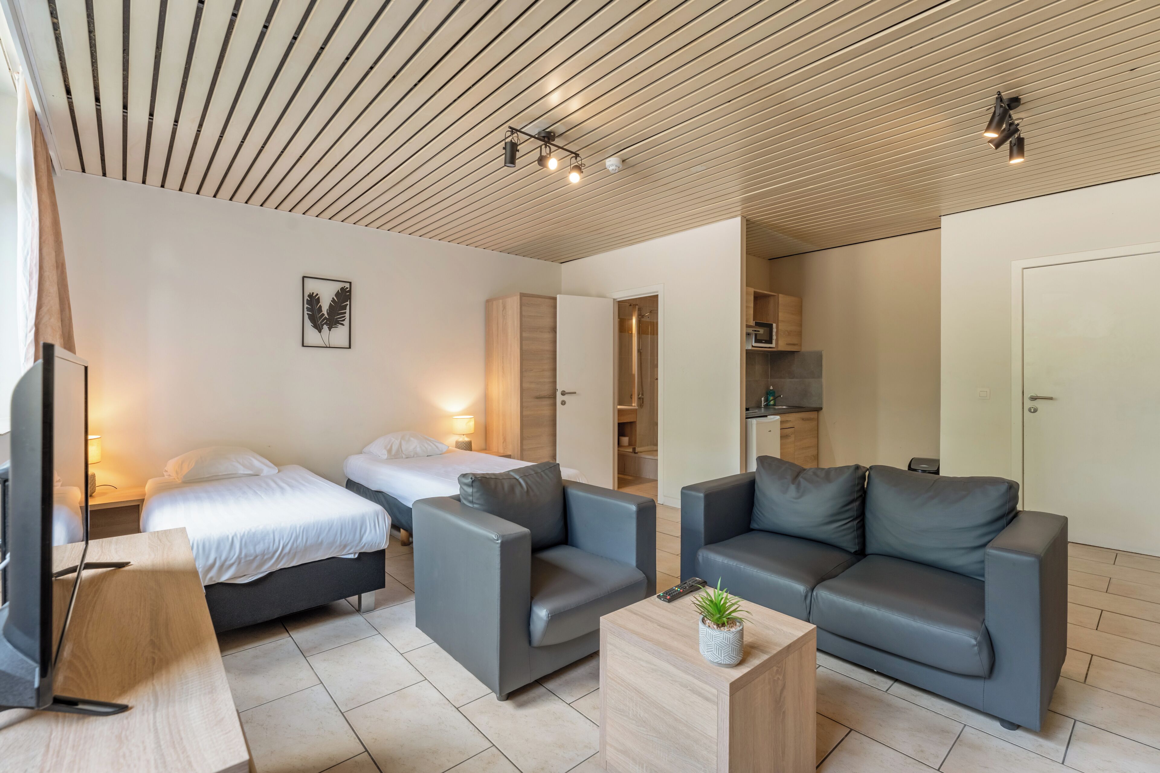 Una habitación de hotel moderna con techo de madera, dos camas, sofás grises, una mesa de centro de madera y un televisor.