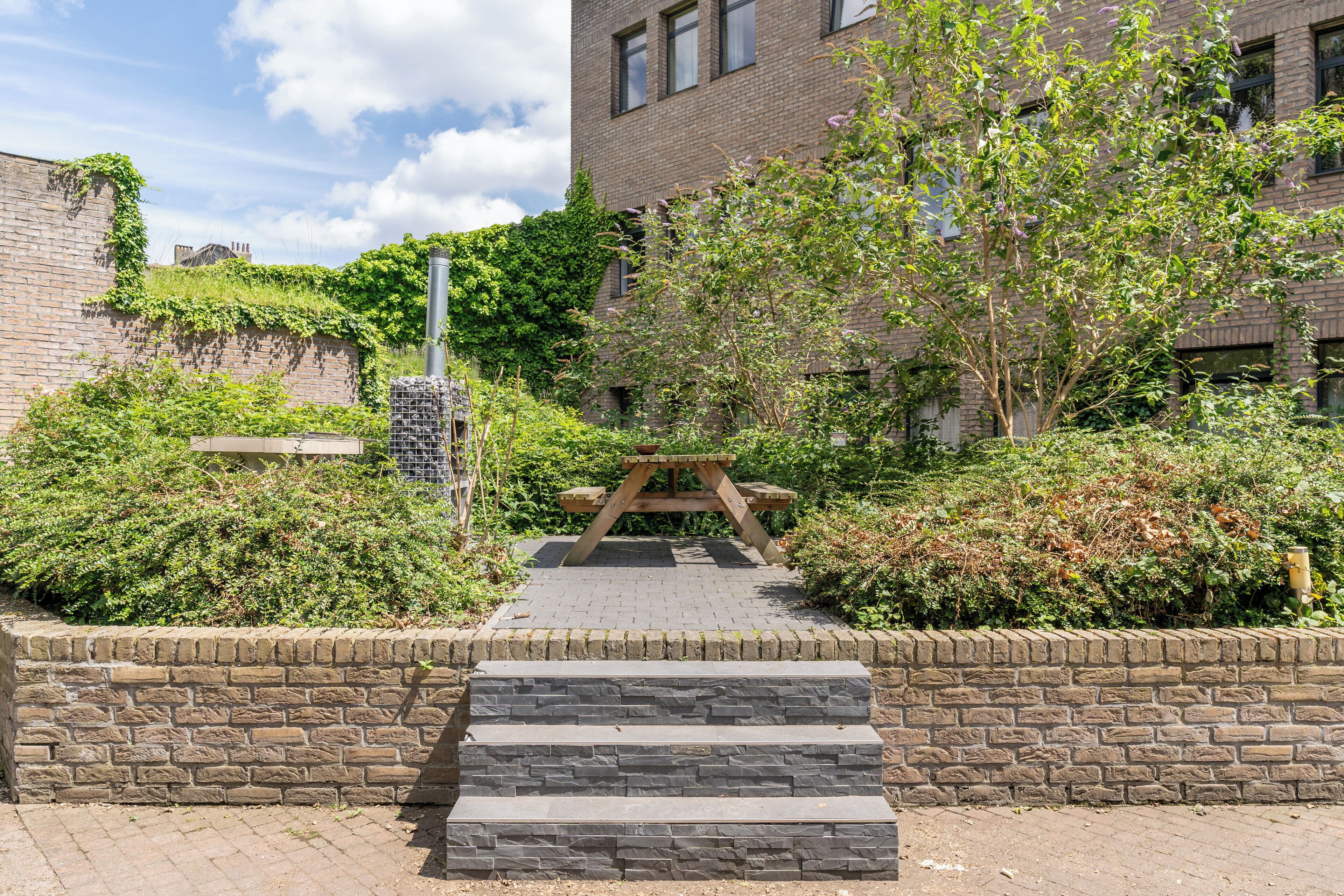 A stone staircase leading to a wooden picnic table surrounded by greenery and a brick building.