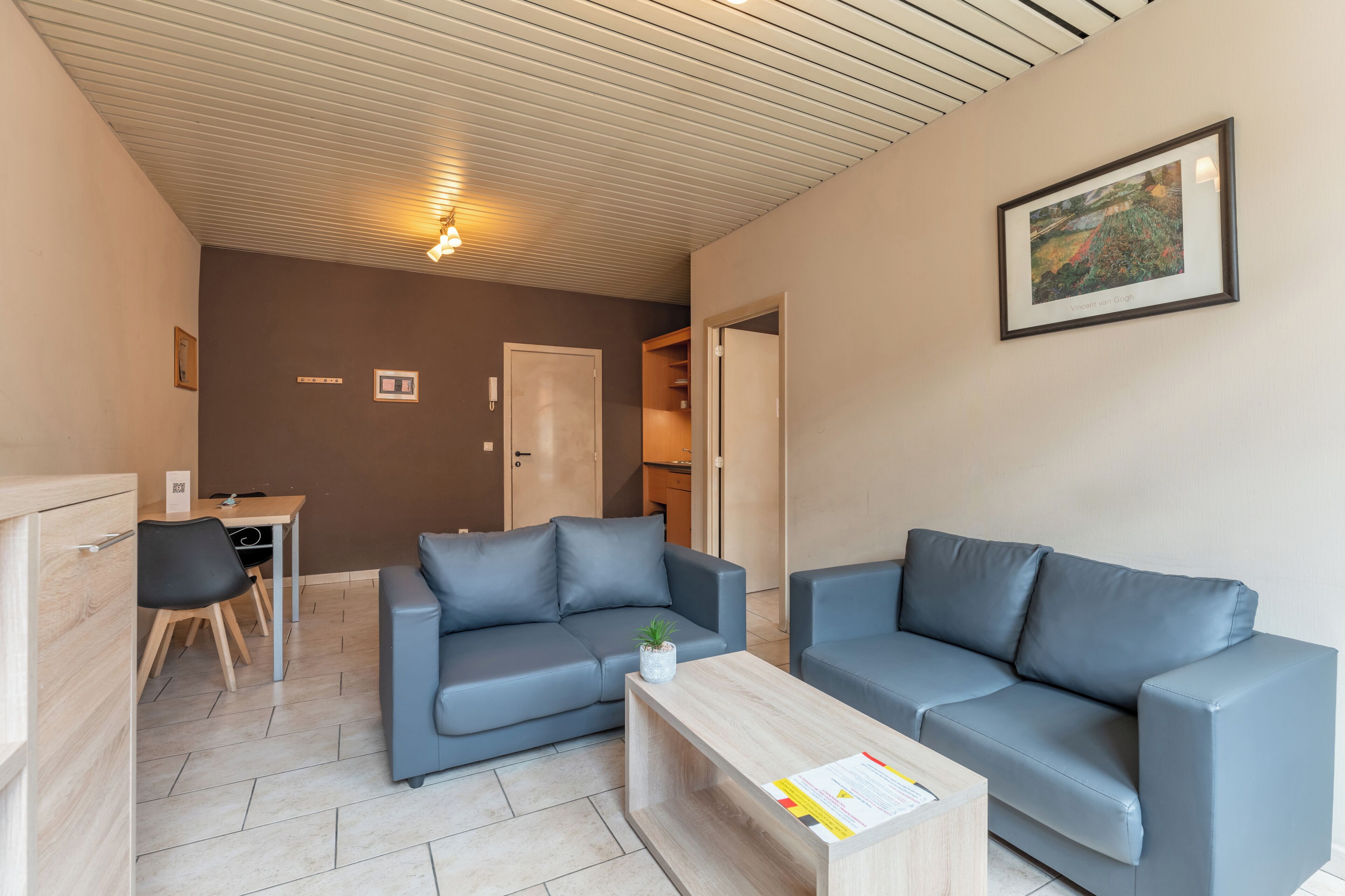A modern living room with two grey sofas, a wooden coffee table, and a small plant. There is a framed artwork on the wall and a door leading to another room.
