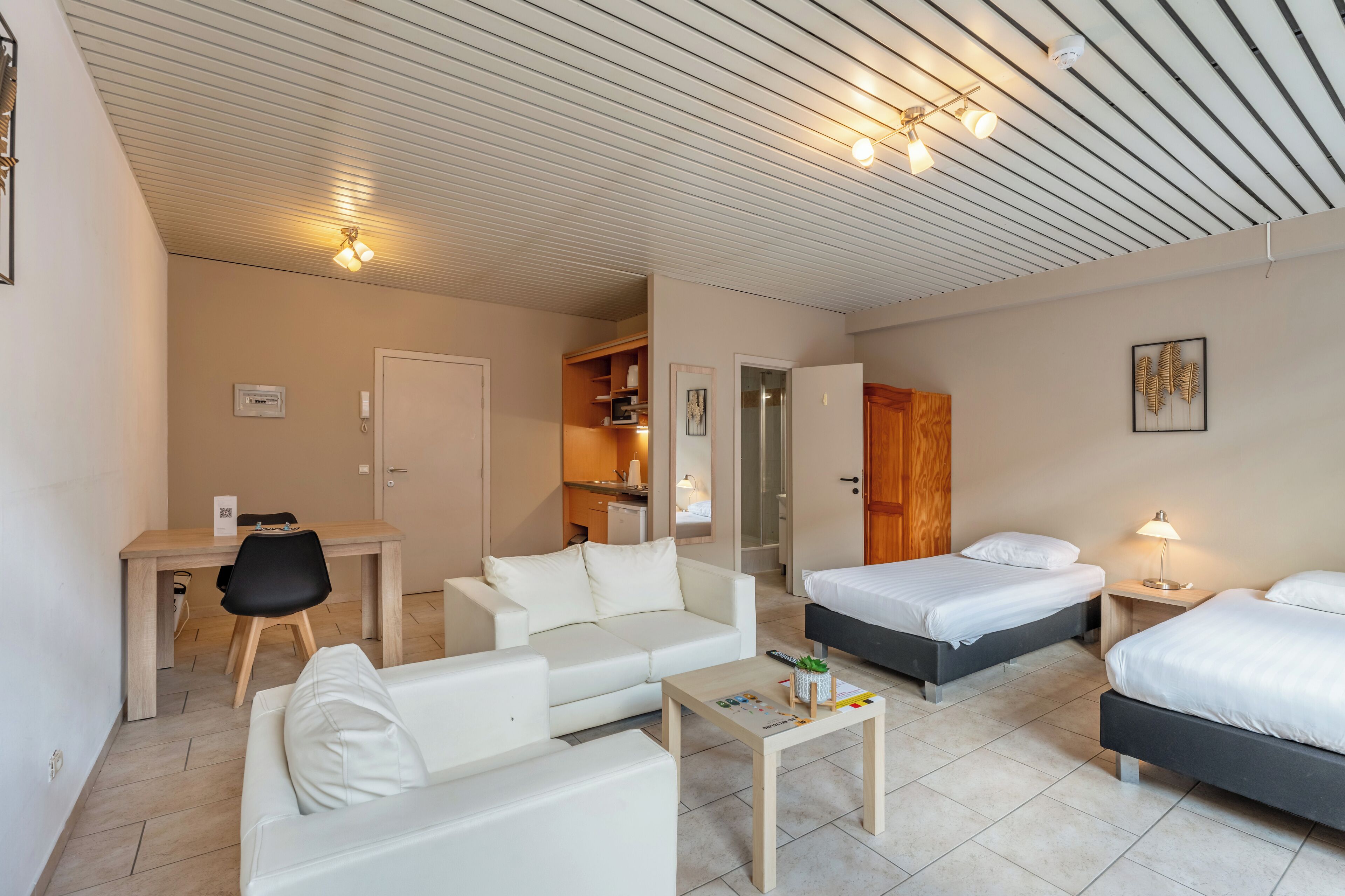 A modern hotel room with a kitchenette, two single beds, a sofa, a desk, and a small table.