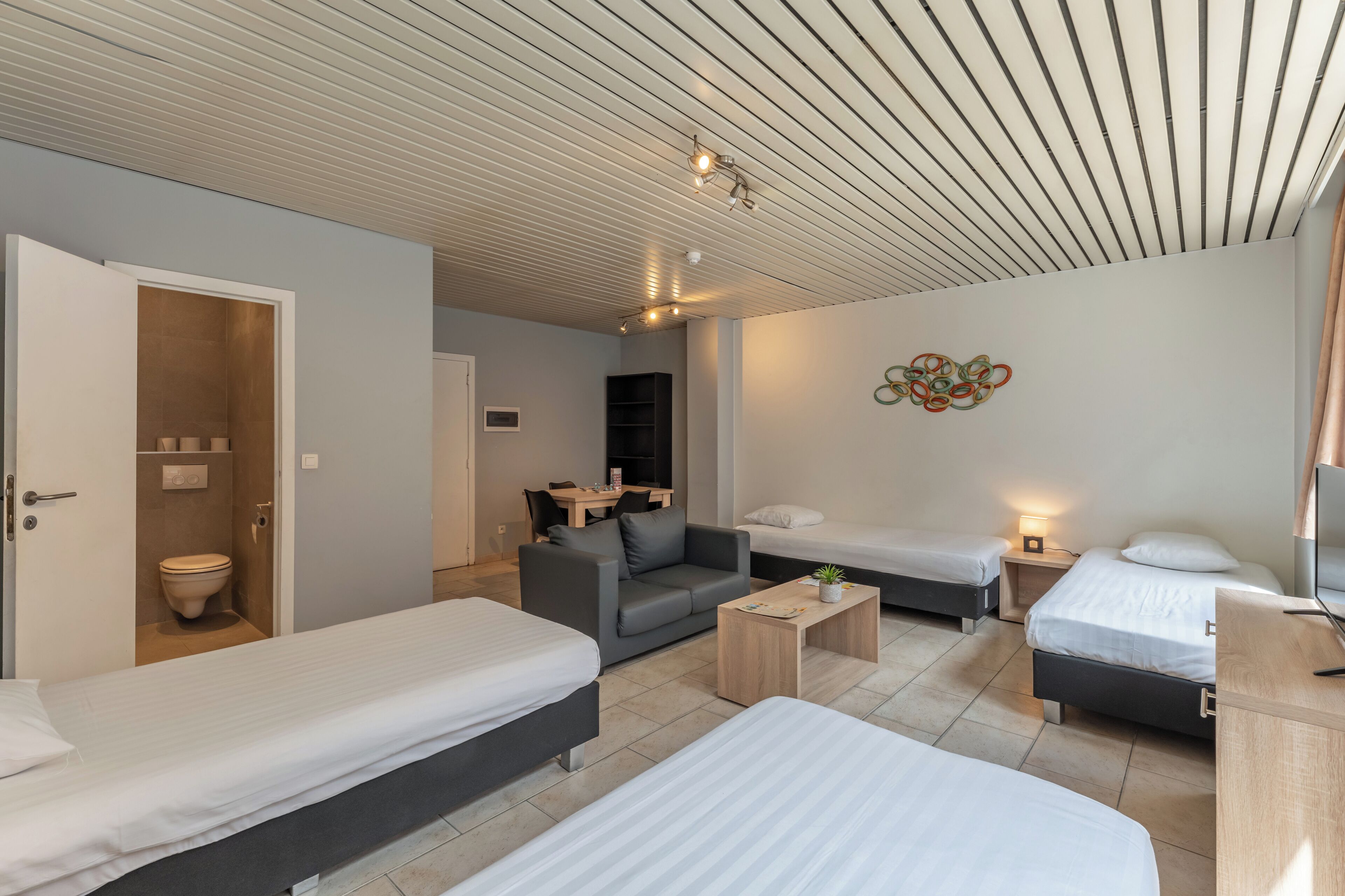 Una habitación moderna y bien iluminada con dos camas individuales, un sofá, una mesita y un baño con inodoro.