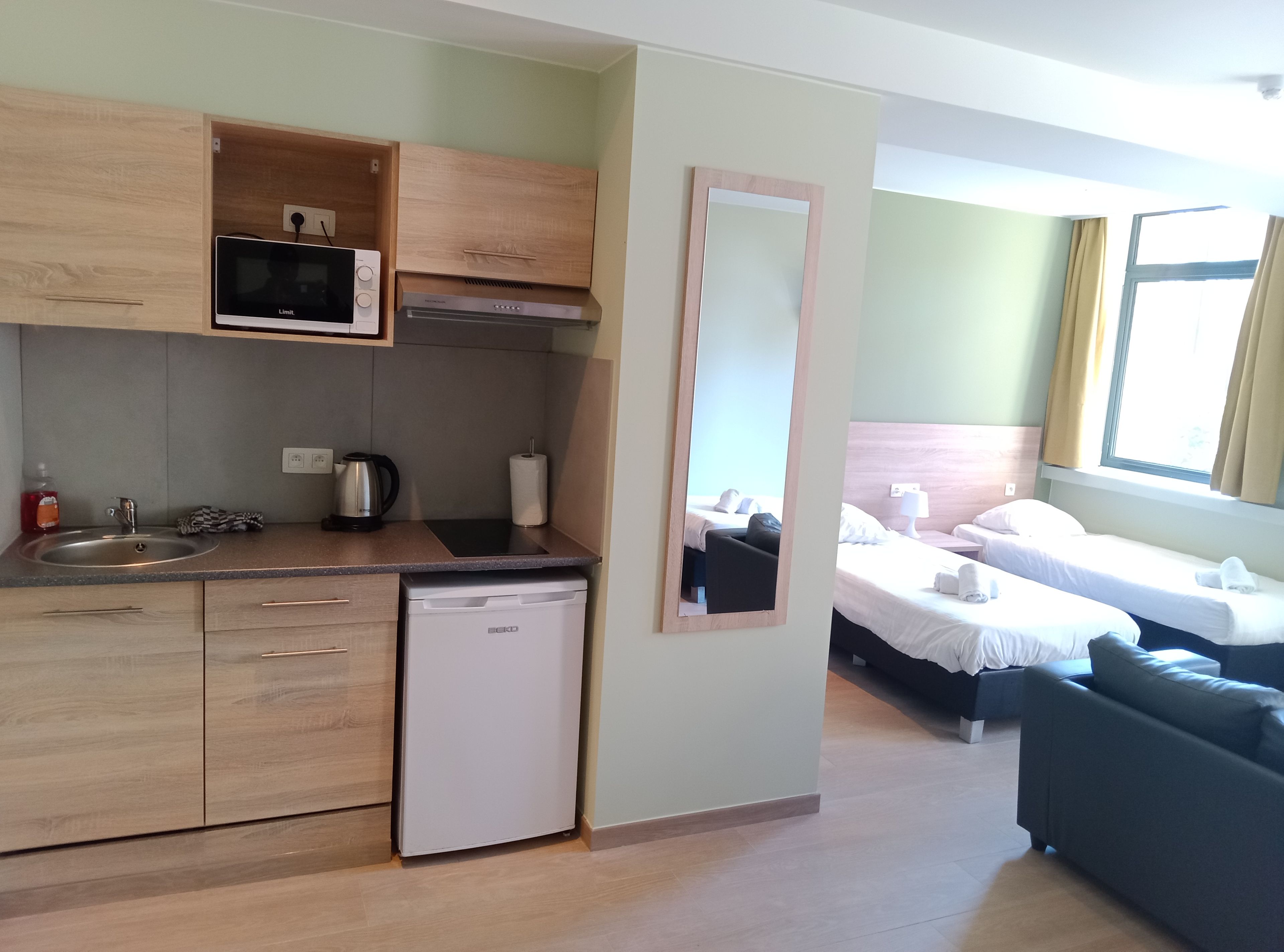 A compact hotel room with a kitchenette, a bed, a sofa, and a large mirror.