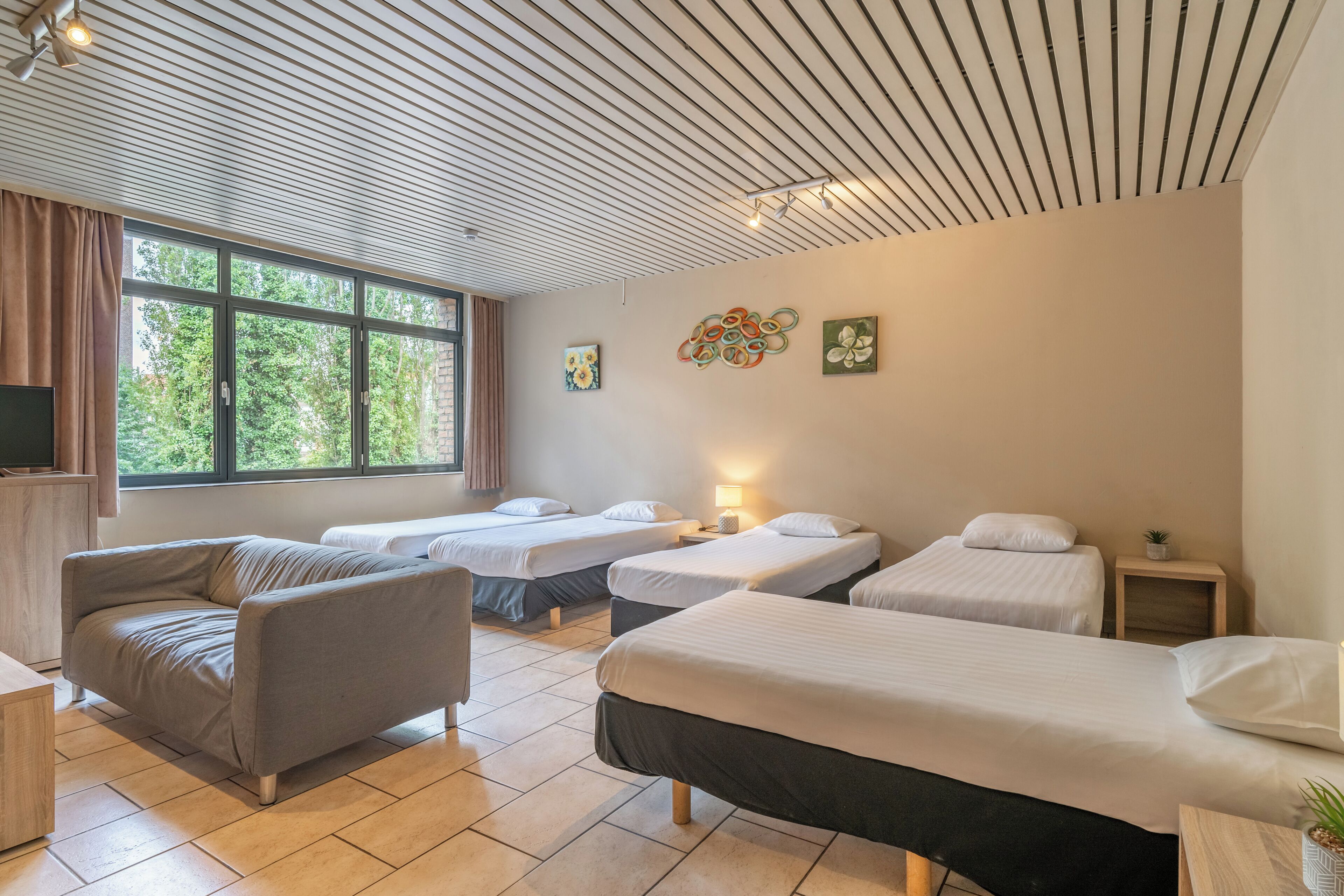 Une chambre d’hôtel avec un canapé, trois lits simples, une télévision et des œuvres d’art accrochées au mur.