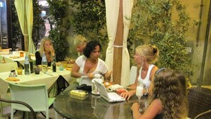 A group of people seated around a round table with a laptop, in an outdoor setting with greenery and a yellow wall in the background. - Hotel Il Ponte (Cecina)