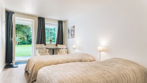 A hotel room with two beds, a small table, two chairs, a window with curtains, and a lamp.