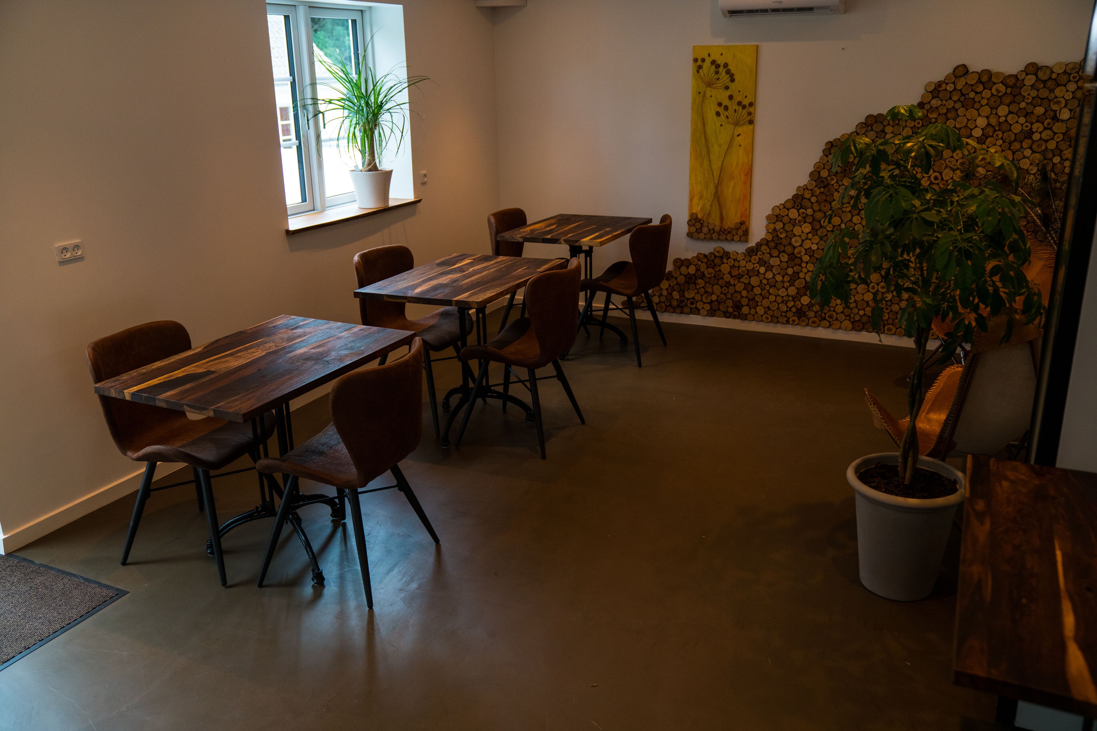 Un espace repas avec des tables et des chaises en bois, une plante en pot et un mur décoré d’un tableau jaune et de piles de bûches.