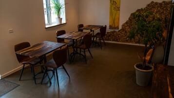 Un espace repas avec des tables et des chaises en bois, une plante en pot et un mur décoré d’un tableau jaune et de piles de bûches.