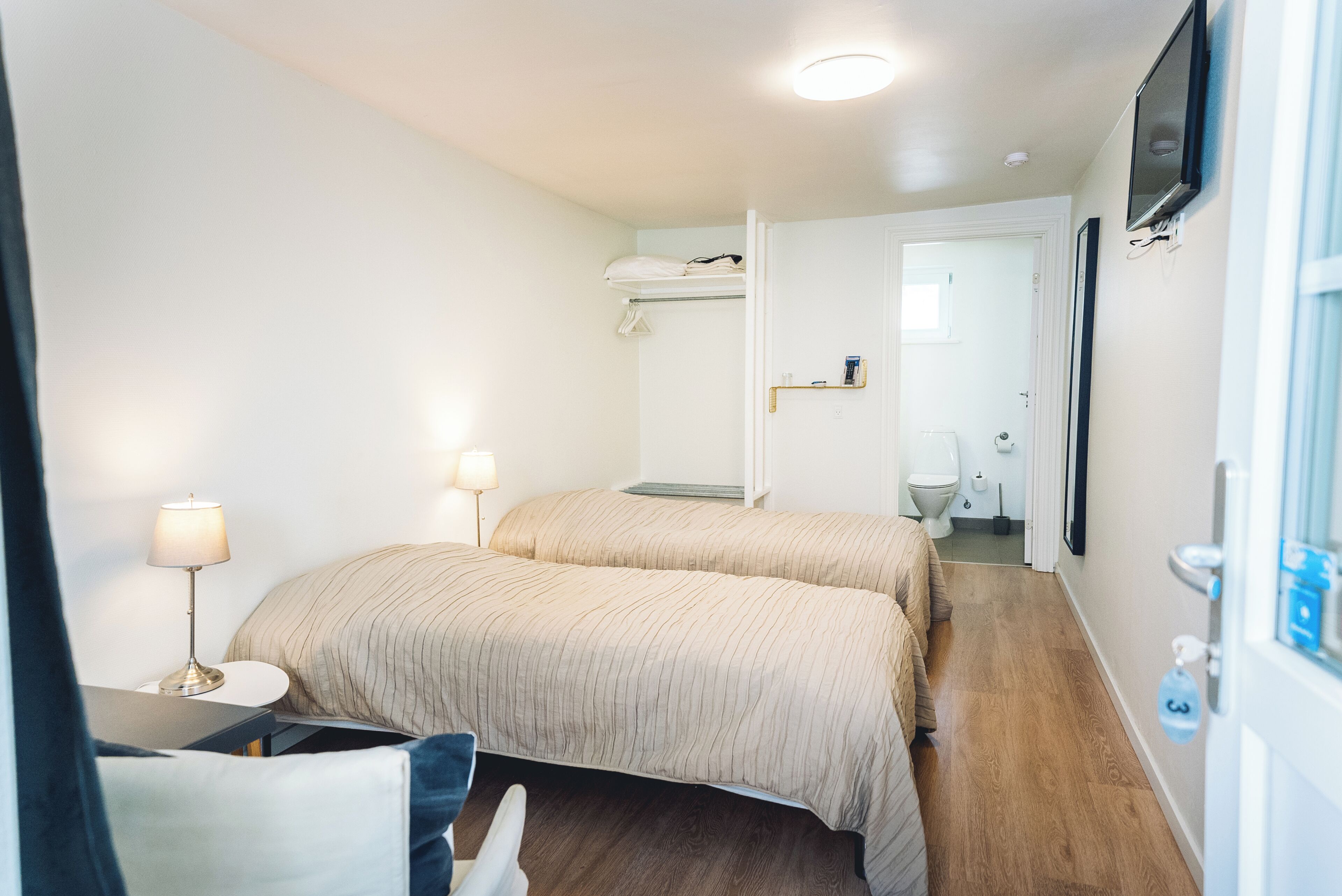 Una habitación de hotel con dos camas, un escritorio con lámpara, un televisor montado en la pared y un baño visible a través de una puerta abierta.