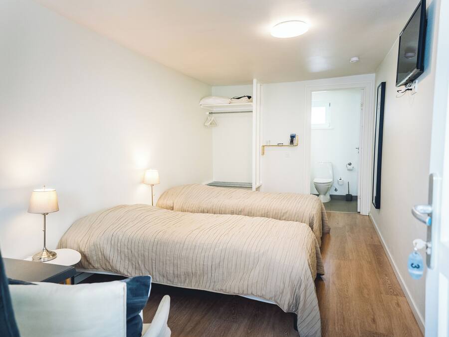 Una habitación de hotel con dos camas, un escritorio con lámpara, un televisor montado en la pared y un baño visible a través de una puerta abierta.