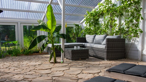 A covered patio area with stone flooring, a wicker sofa with cushions, and a potted plant.