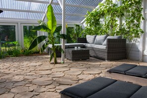 Ein überdachter Patio-Bereich mit Steinboden, einem Rattangeflechtsofa mit Kissen und einem Blumentopf.