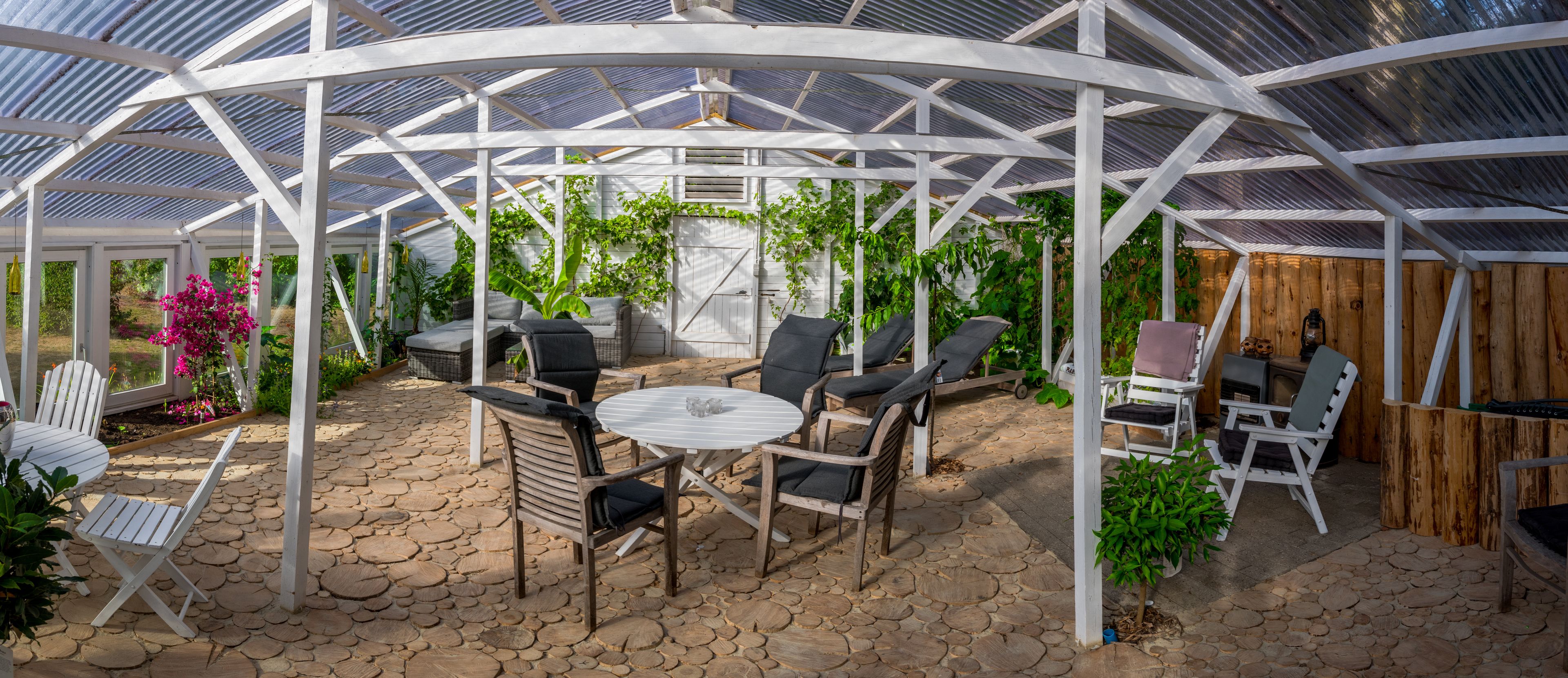Ein überdachter Patio-Bereich mit Steinböden, weißen Holzkufen, einem runden Tisch und Blumentöpfen.
