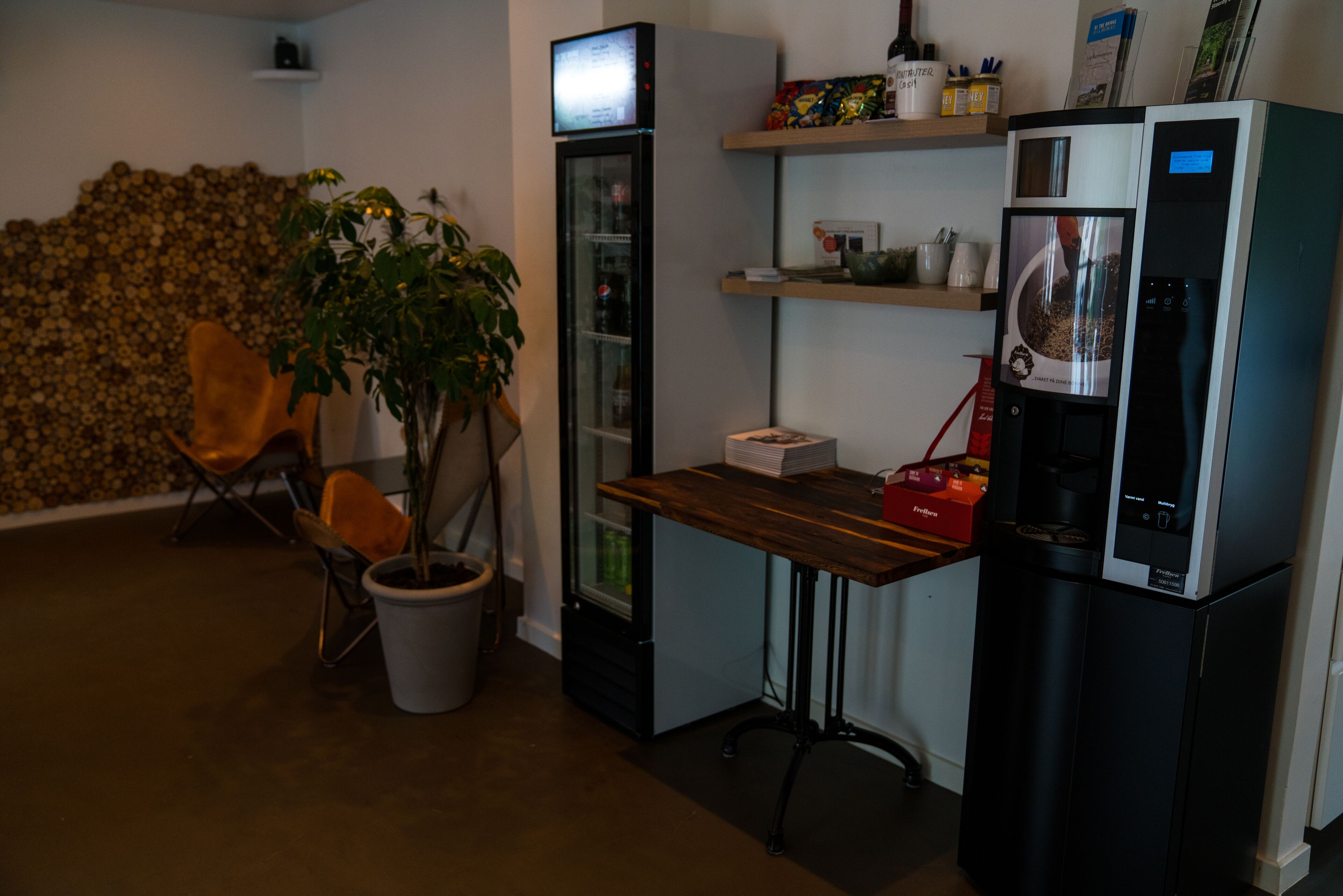 A compact kitchenette with a refrigerator, microwave, and coffee maker. There is a wooden table, a potted plant, and a chair.