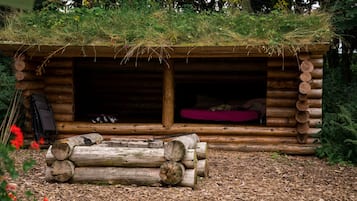 A rustic log cabin with a thatched roof, a sleeping area with a red cushion, and a pile of logs in front.