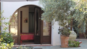 Un espace extérieur aménagé avec une table et des chaises, une grande plante en pot et une porte donnant sur une pièce où se trouve un lit recouvert d’une courtepointe rouge.
