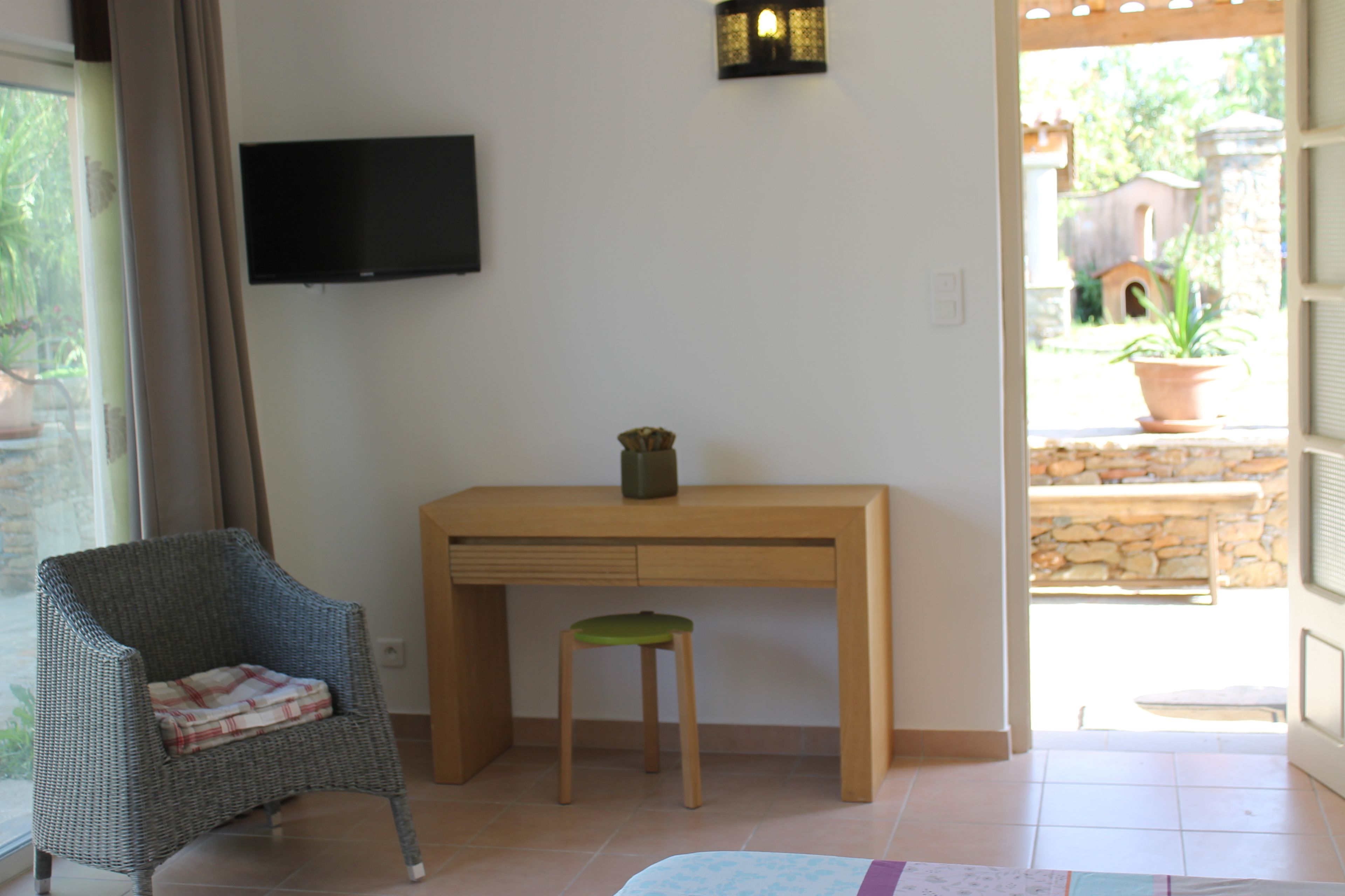 A room with a wooden desk, a wicker chair, a small stool, a television mounted on the wall, and a view of the outside through a glass door.