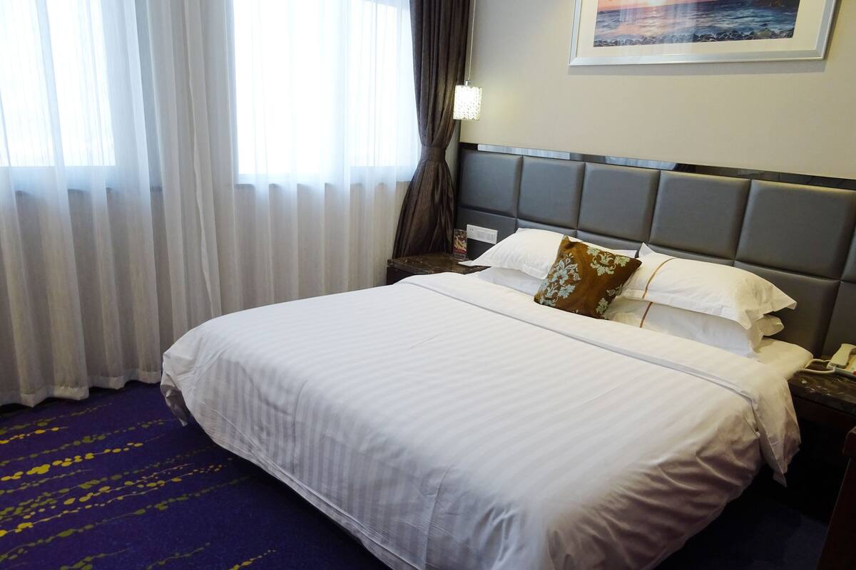 Ein Hotelzimmer mit einem großen Bett, weißer Bettwäsche, einem Kopfteil mit einer Leuchte, einem Nachttisch mit einem Telefon und einem Fenster mit durchsichtigen Vorhängen.