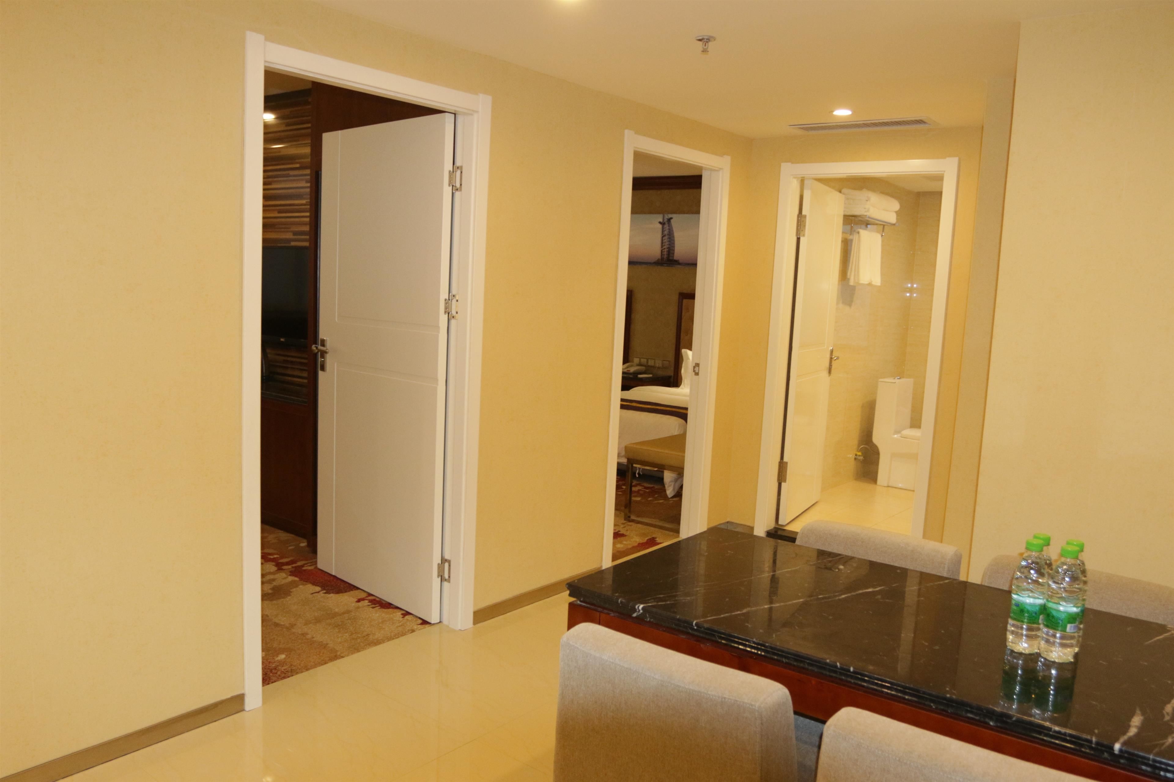 Habitación de hotel con zona de comedor que incluye una mesa de mármol y dos sillas, baño con ducha de vidrio y pasillo con puerta que da a otra habitación.