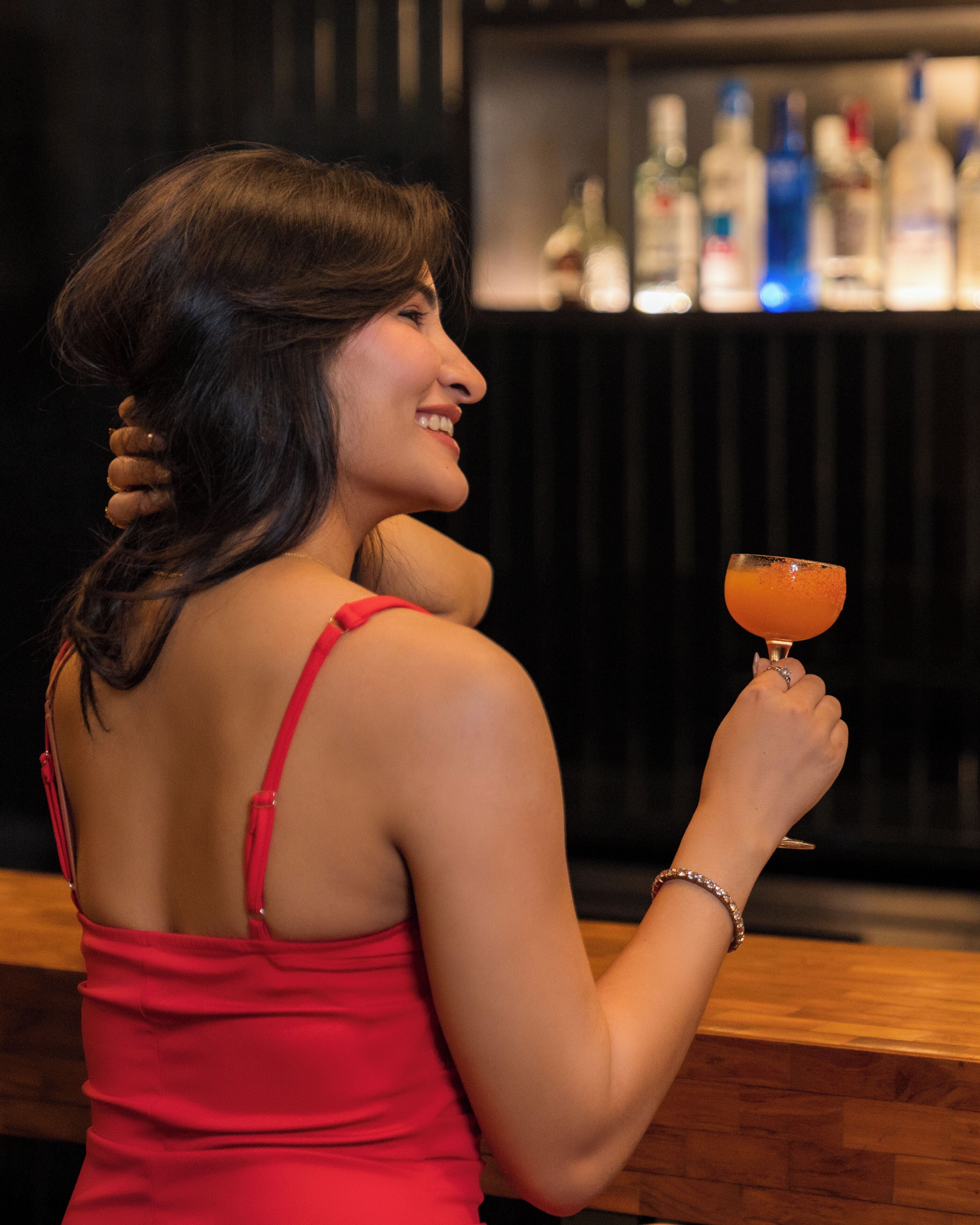 A person holding a cocktail glass, dressed in a red dress, with a bar and bottles in the background.