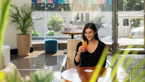 A person sitting at a table with a drink, in a room with large windows and potted plants. - Park Inn by Radisson New Delhi IP Extension (New Delhi)