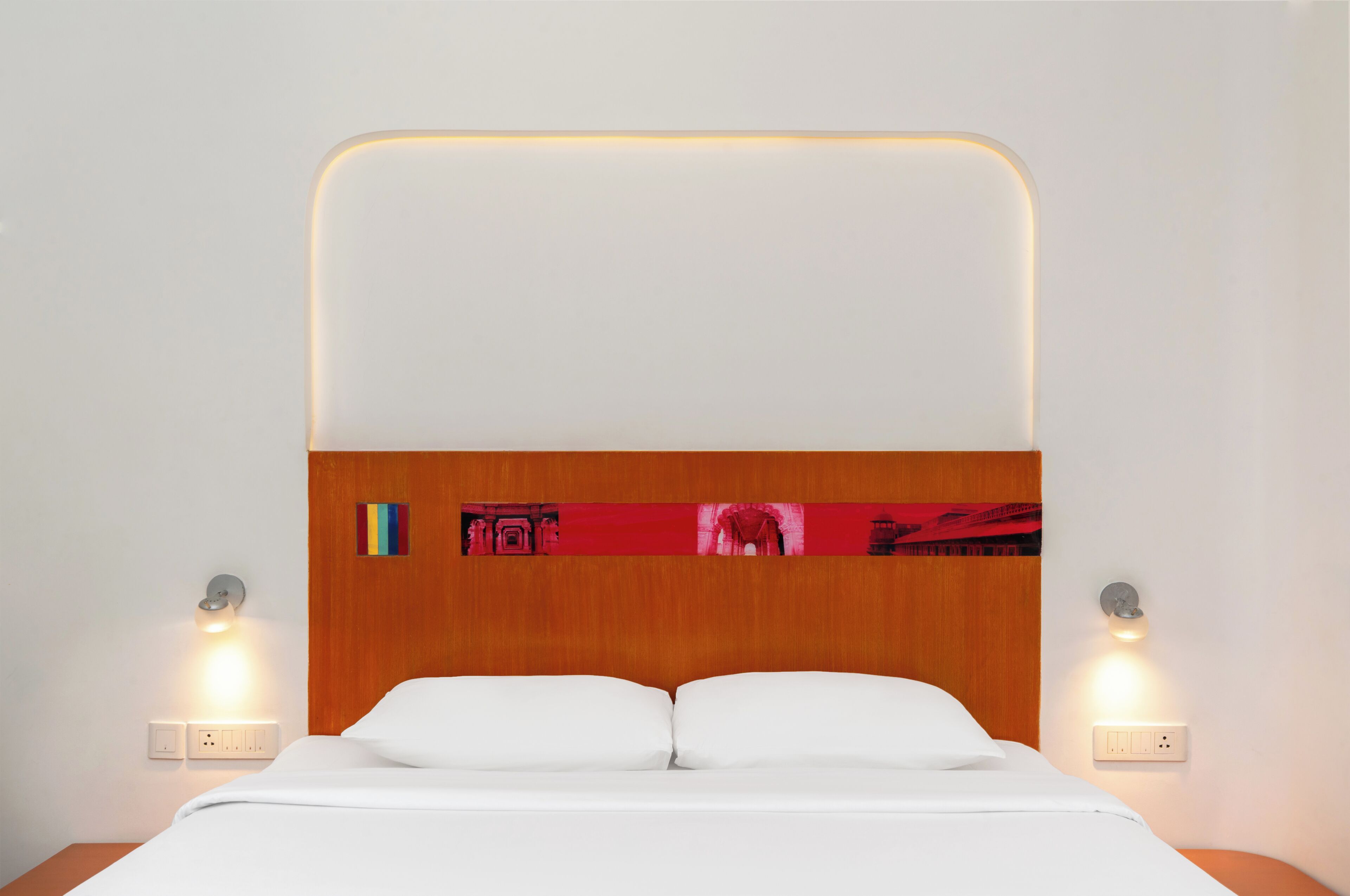 Une chambre d’hôtel avec une tête de lit en bois, deux lampes de chevet, un lit avec du linge de maison blanc et une œuvre d’art accrochée au mur.