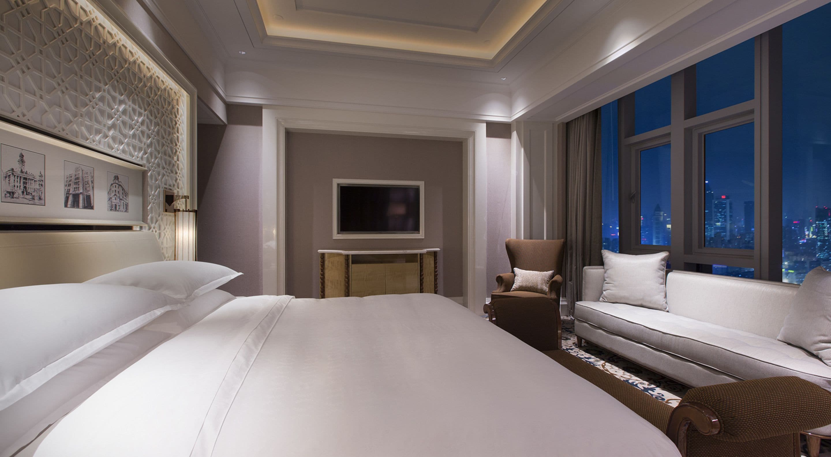 A modern hotel room with a large bed, a sofa, a TV, and a city view.