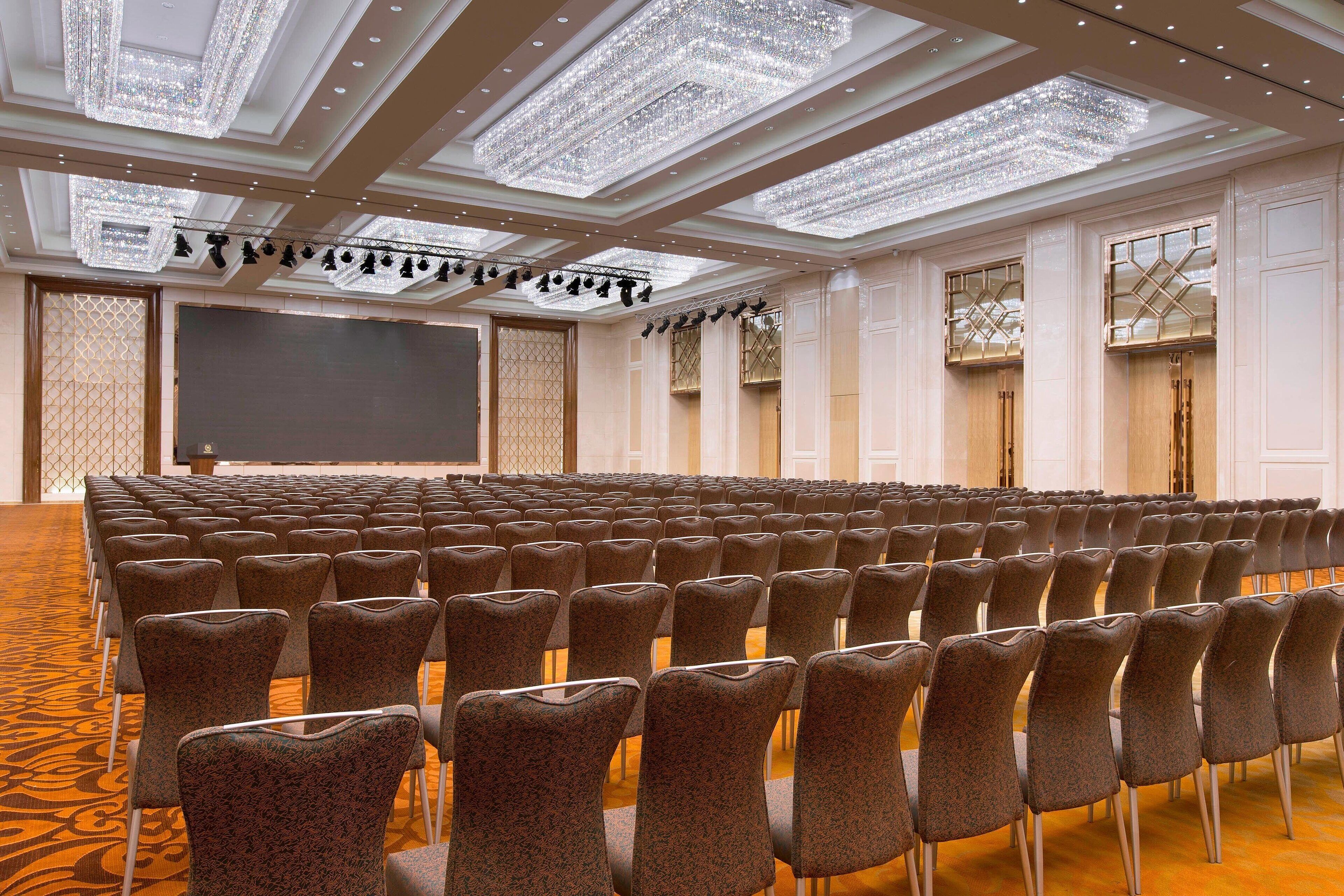 Un salón de conferencias con filas de sillas, una pantalla grande y lámparas de techo decorativas.