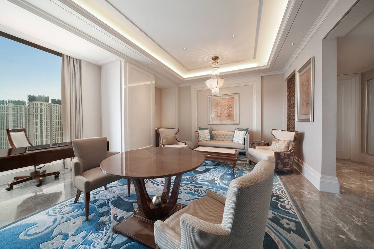 Een ruime kamer met een ronde tafel, comfortabele zithoek, een piano en een groot raam met uitzicht op de stad.