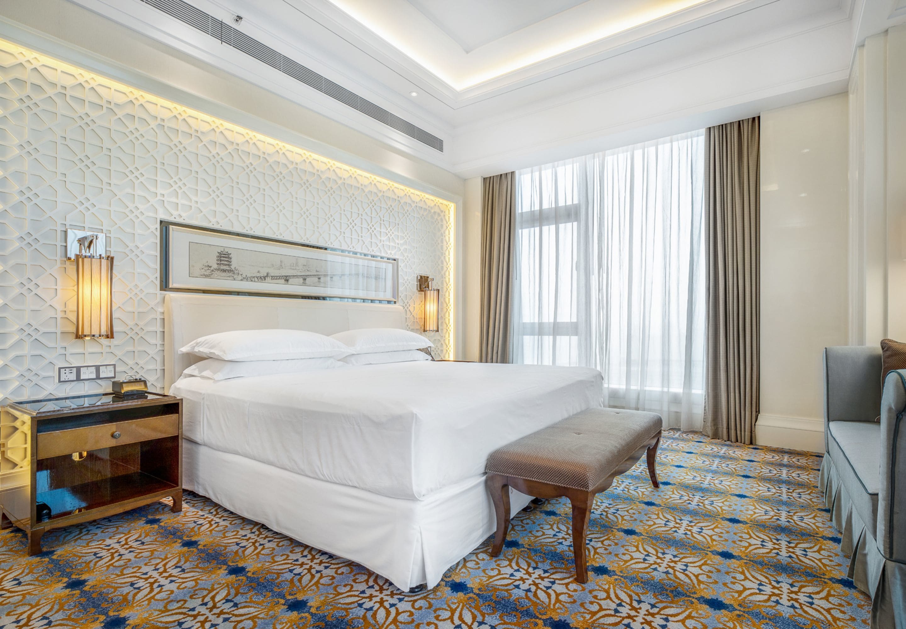 A hotel room with a large bed, a bedside table, a bench, a TV, and a patterned carpet.