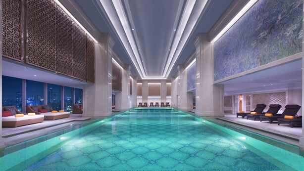 A large indoor swimming pool with a geometric tile pattern, lounge chairs, and seating areas with cushions.