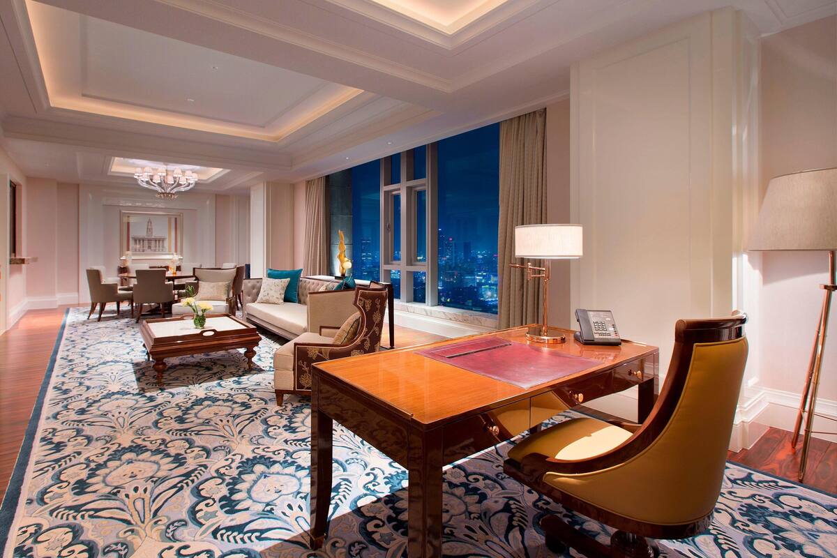 Ruime hotelkamer met een groot raam en uitzicht op de stad, een zithoek met een tafel en stoelen, en een zangle plek met een bank en fauteuils.