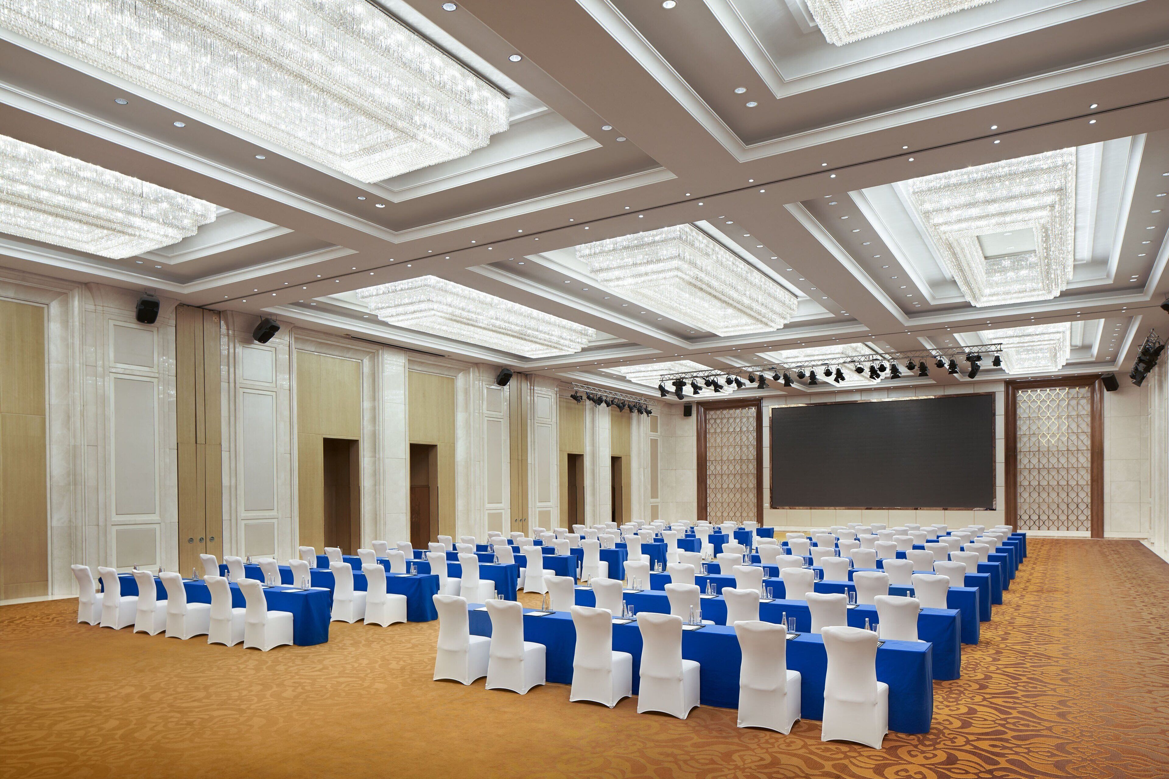 Un amplio salón de conferencias con filas de mesas y sillas, una pantalla grande y un techo alto con candelabros.