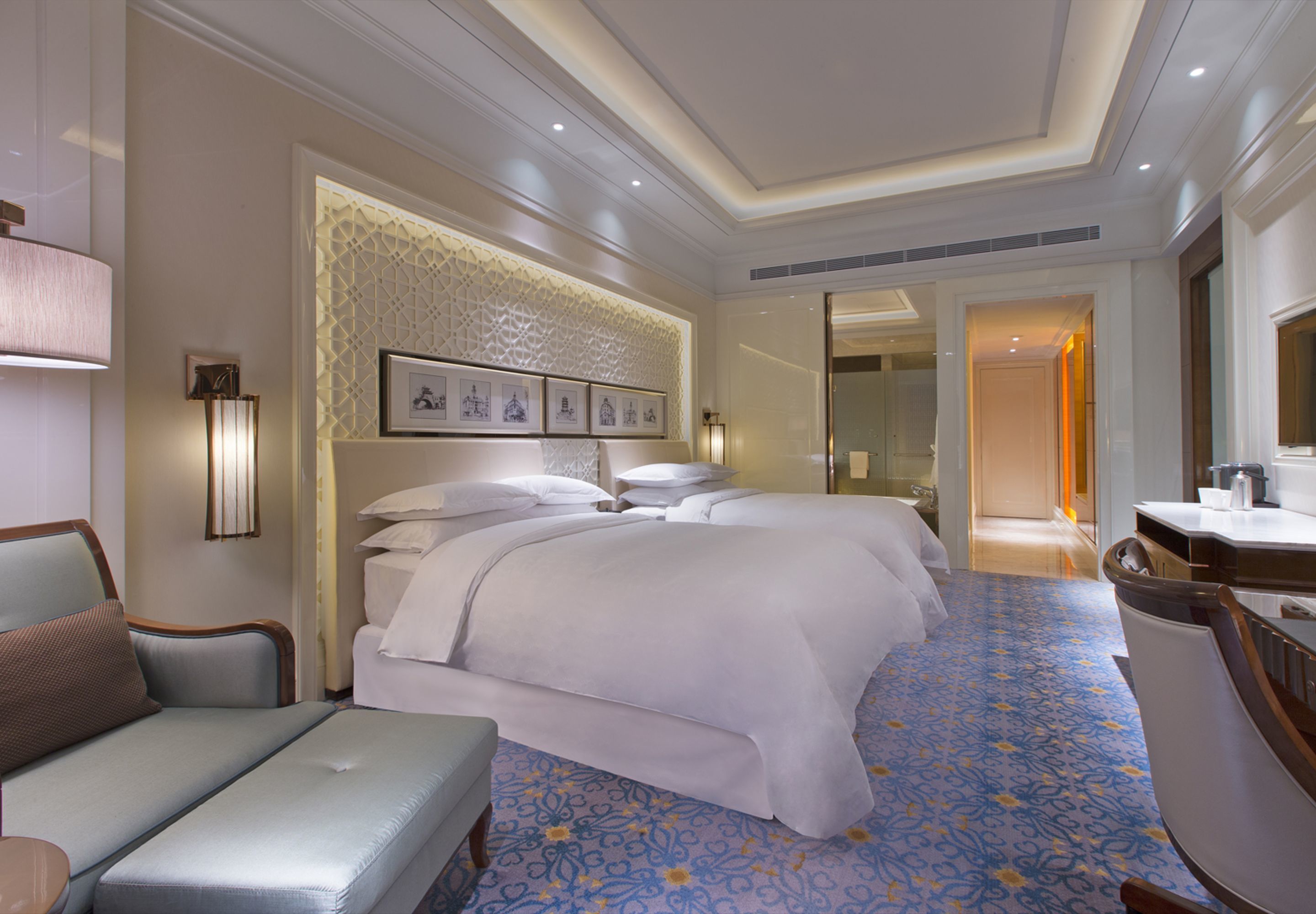 A hotel room with a large bed, a desk, and a chair. There is a wall with framed pictures and a decorative panel.