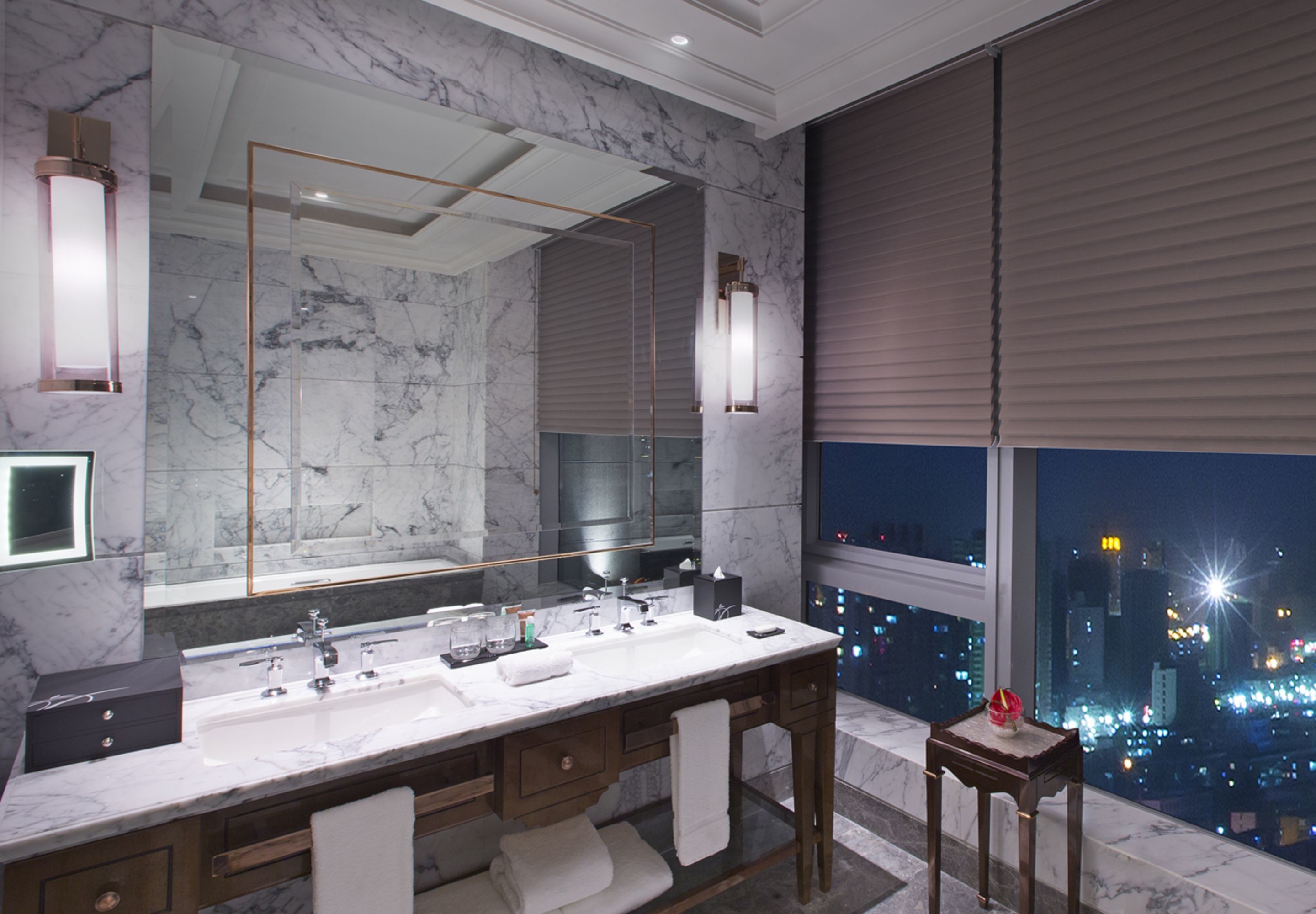 A modern bathroom with a large mirror, dual sinks, and a view of the city at night.