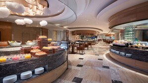 A modern hotel lobby with a marble reception desk, buffet area with various food items, and a seating area with tables and chairs.