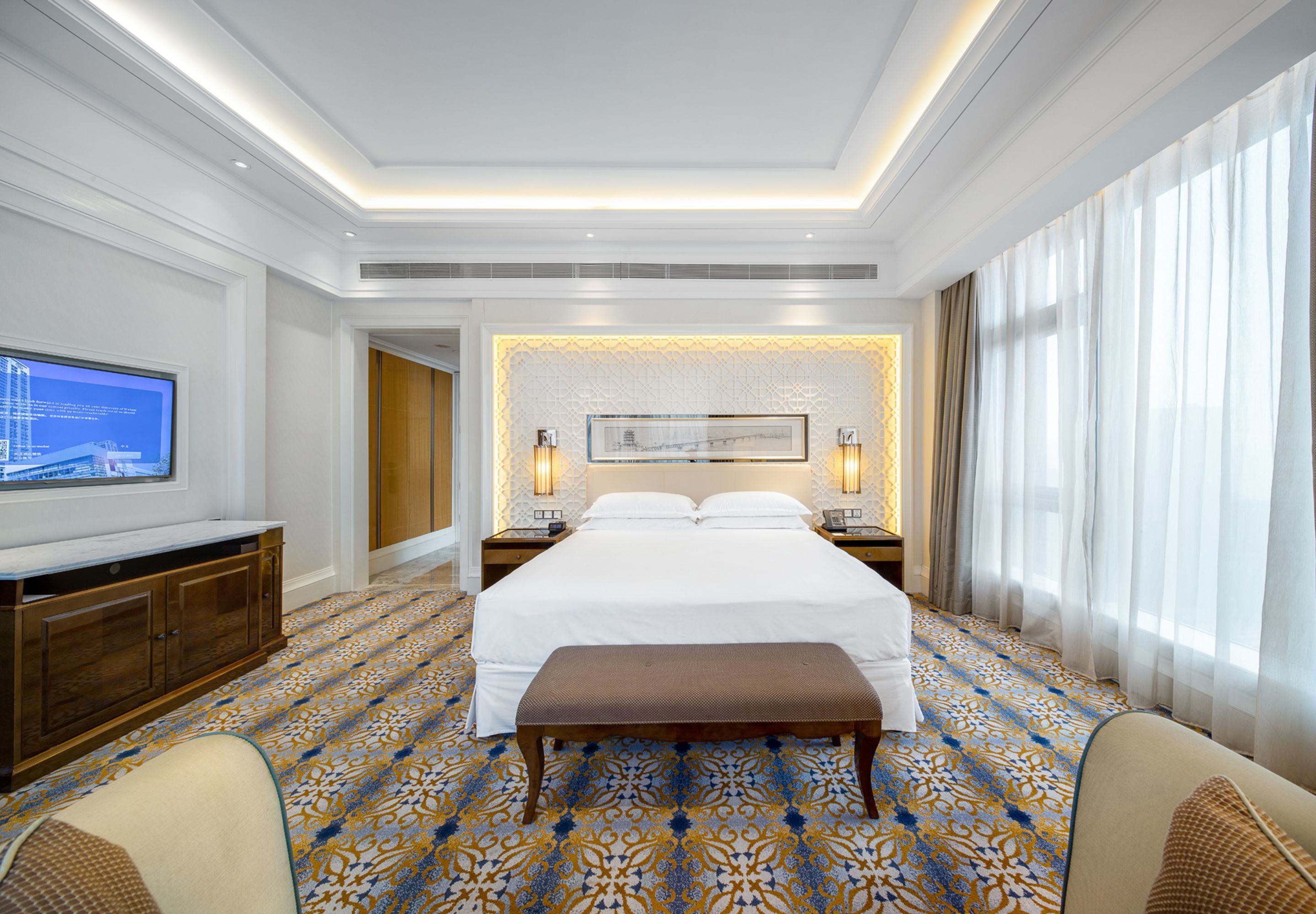 A hotel room with a large bed, a television on the left, a wooden bench, and a patterned carpet.