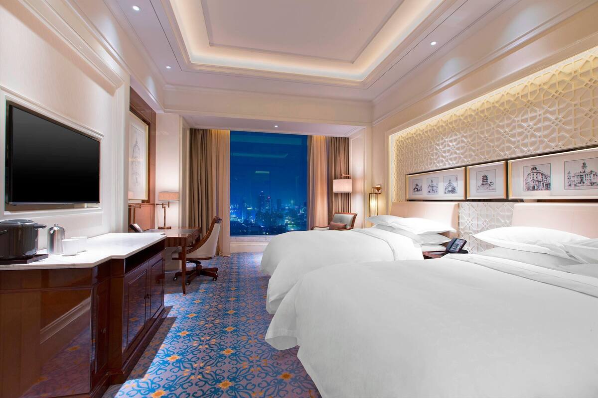 Hotelkamer met twee bedden, een bureau, een flatscreen televisie en uitzicht op de stad 's nachts, met een kamerbreed tapijt.
