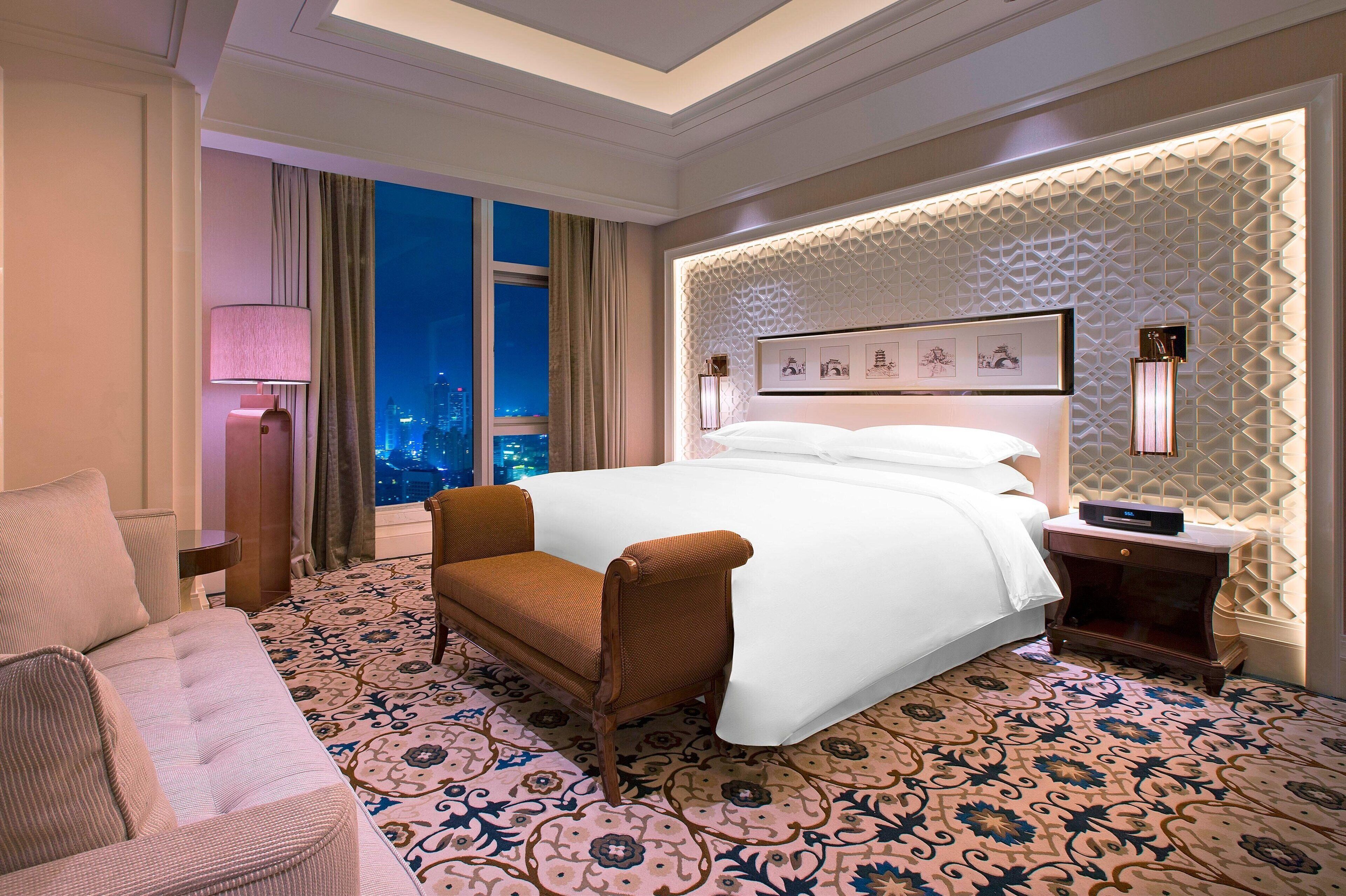 A hotel room with a large bed, a sofa, a nightstand, and a view of the city at night.