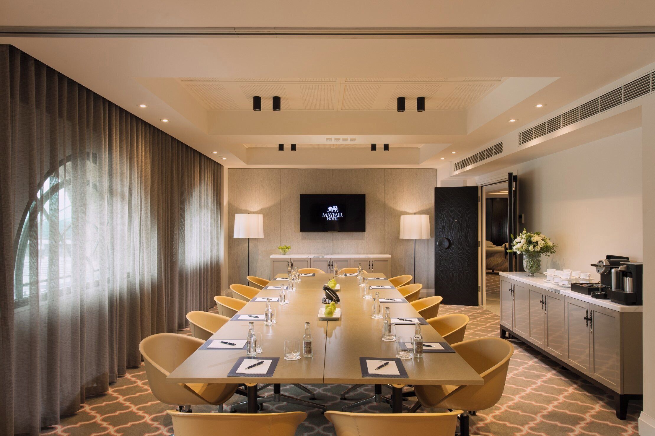 A conference room with a long table, chairs, a TV screen, and a kitchenette area.